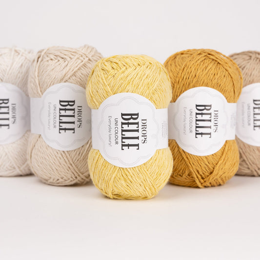 Fünf Knäuel der Wolle DROPS Belle in den Farben 26 perle, 03 hellbeige, 04 löwenzahn, 37 ocker und 28 keksteig (von links nach rechts)