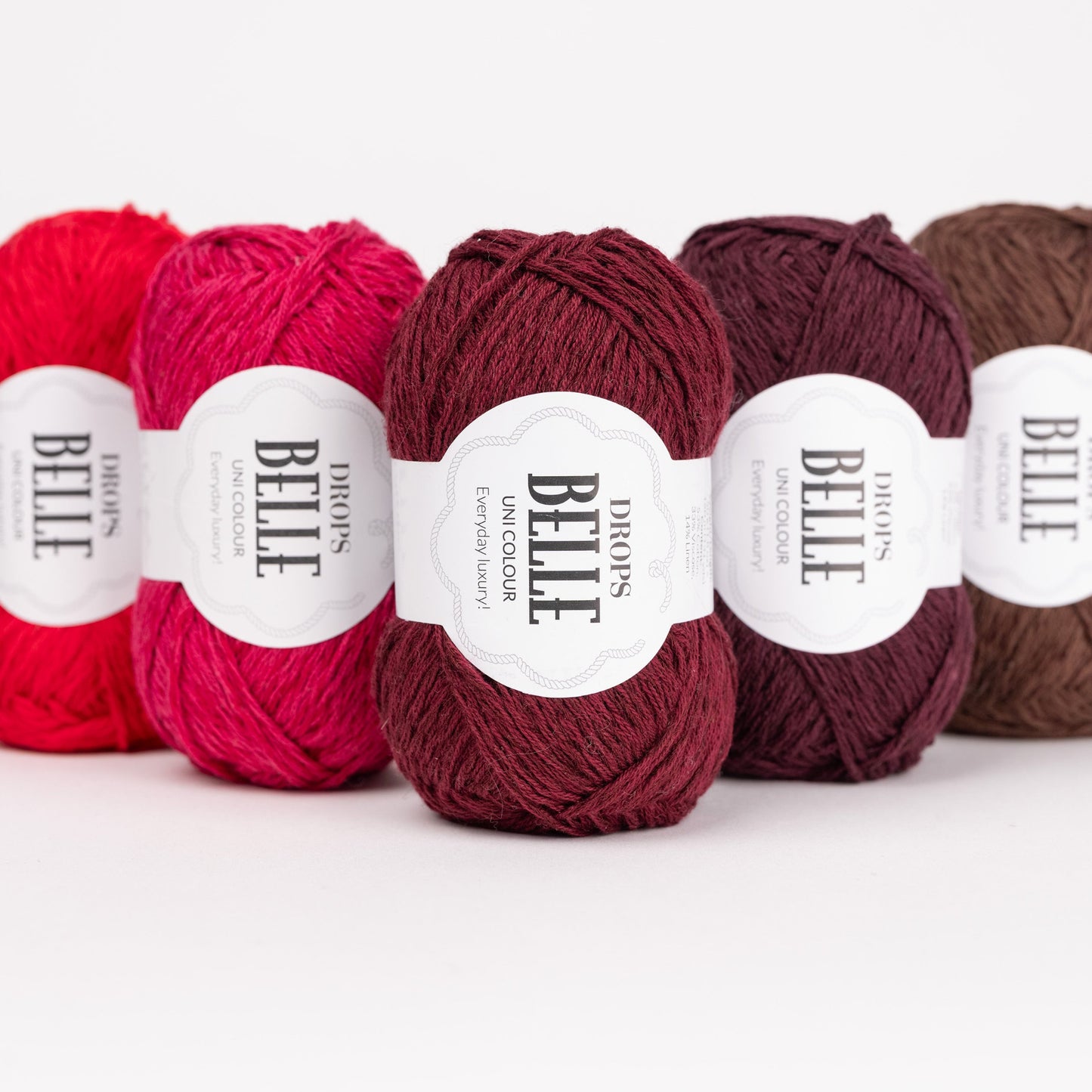 Fünf Knäuel der Wolle DROPS Belle in den Farben 29 rot, 12 kirsche, 36 rubinrot, 40 pflaumenwein und 39 schokolade (von links nach rechts).