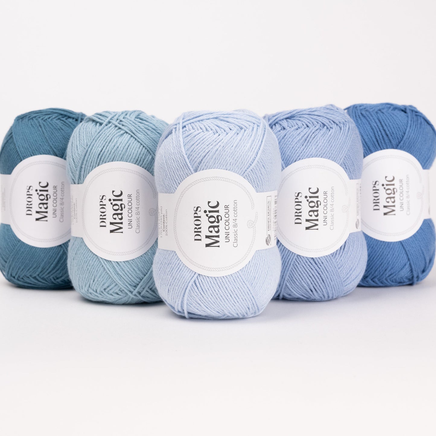 Fünf Knäuel der Wolle DROPS Magic in den Farben 74 blauer hafen, 77 meeresnebel, 26 eisblau, 43 hell jeansblau und 07 jeansblau (von links nach rechts).