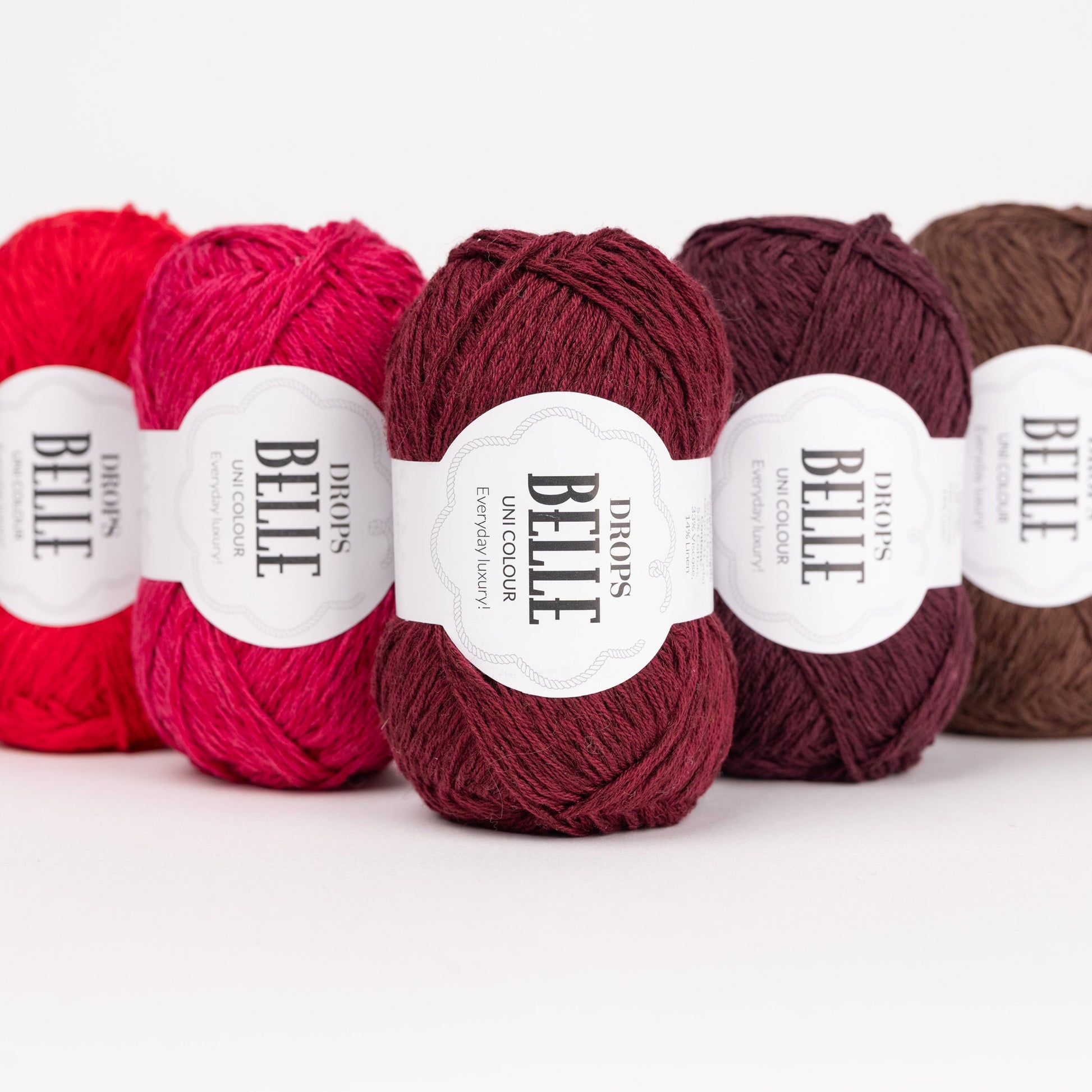 Fünf Knäuel der Wolle DROPS Belle in den Farben 29 rot, 12 kirsche, 36 rubinrot, 40 pflaumenwein und 39 schokolade (von links nach rechts). 