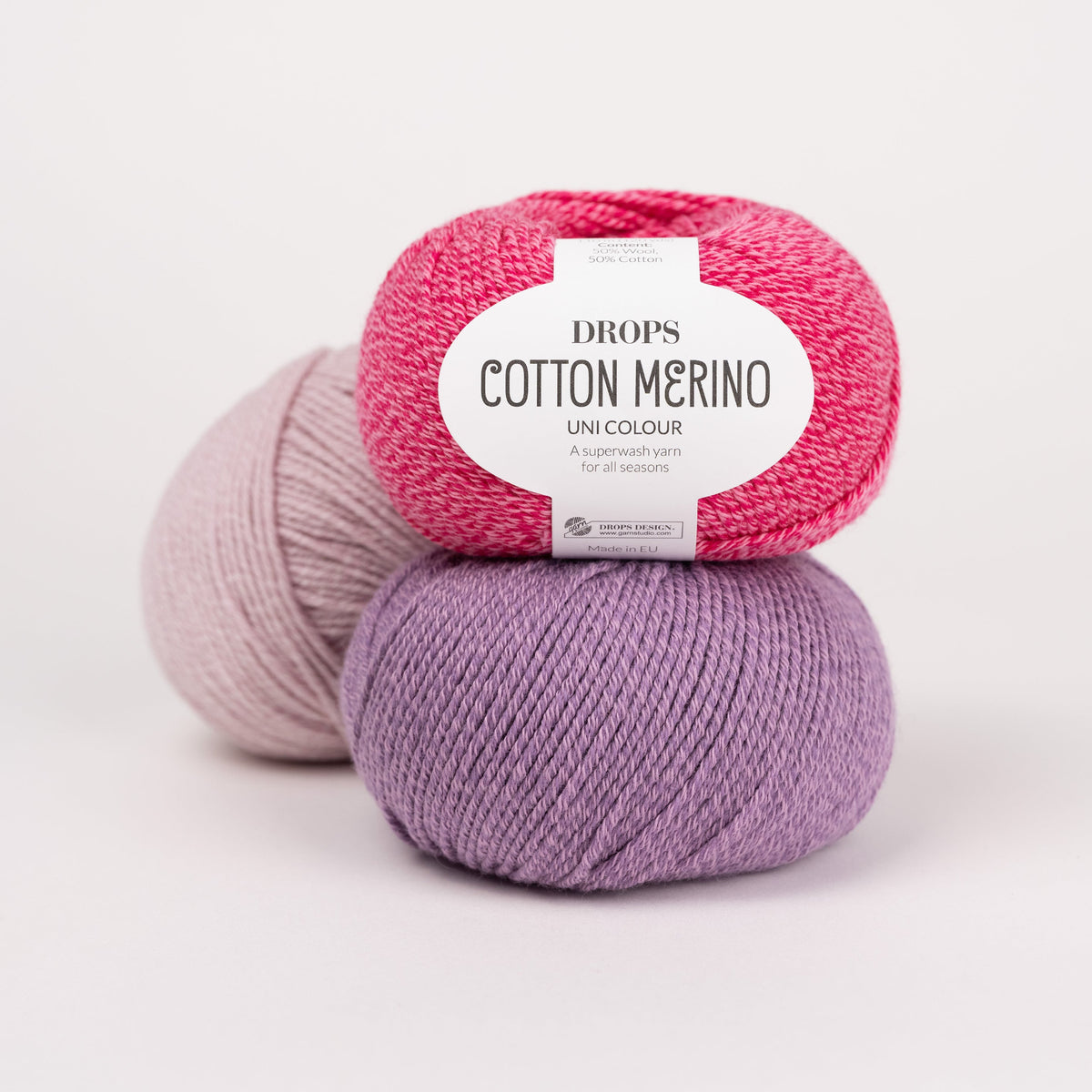 Drei Knäuel der Wolle DROPS Cotton Merino in den Farben 36 himbeersorbet (oben), 23 lavendel (unten) und 05 puderrosa (hinten).