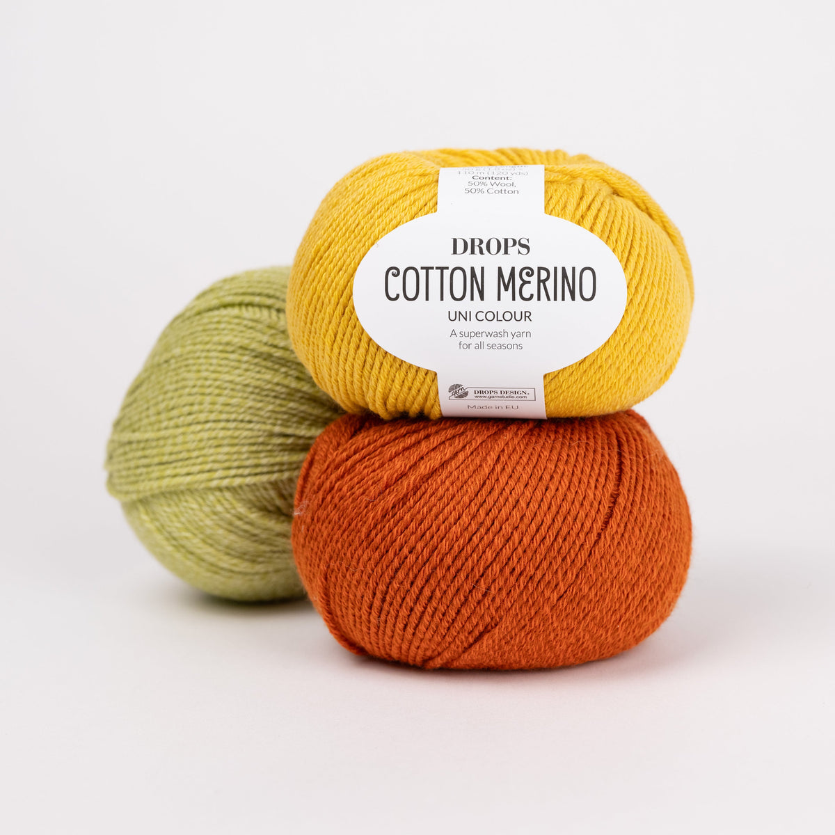 Drei Knäuel der Wolle DROPS Cotton Merino in den Farben 15 senf (oben), 25 rost (unten) und 10 pistazie (hinten).
