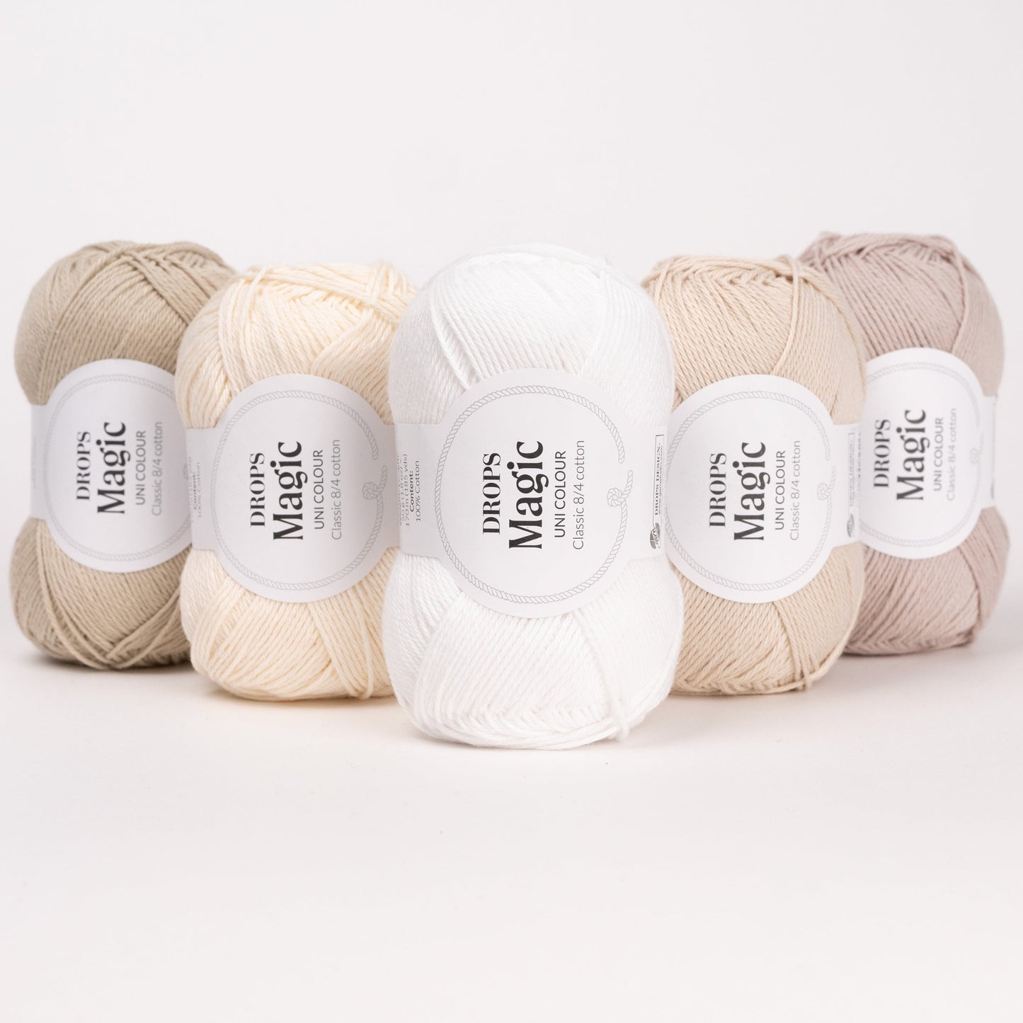 Fünf Knäuel der Wolle DROPS Magic in den Farben 36 tau, 70 natur, 01 weiss, 35 hellbeige und 03 sandperle (von links nach rechts).