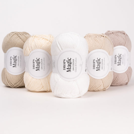 Fünf Knäuel der Wolle DROPS Magic in den Farben 36 tau, 70 natur, 01 weiss, 35 hellbeige und 03 sandperle (von links nach rechts).