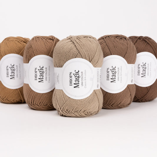 Fünf Knäuel der Wolle DROPS Magic in den Farben 59 karamell, 60 mandel, 61 nougat, 37 wallnuss und 49 kaffee (von links nach rechts).