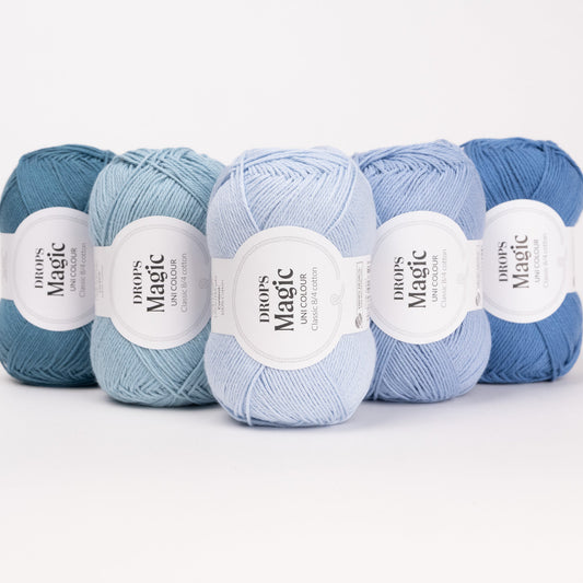 Fünf Knäuel der Wolle DROPS Magic in den Farben 74 blauer hafen, 77 meeresnebel, 26 eisblau, 43 hell jeansblau und 07 jeansblau (von links nach rechts).