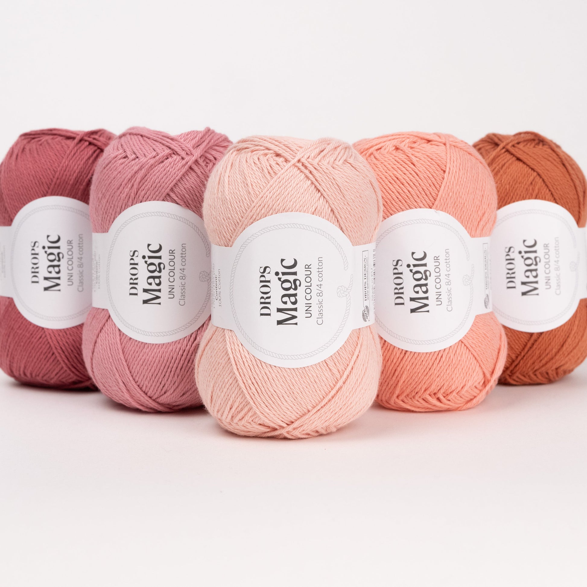 Fünf Knäuel der Wolle DROPS Magic in den Farben 79 dunkle rose, 22 zarte rose, 71 hell pfirsich, 54 pfirsich und 34 rostrot (von links nach rechts).