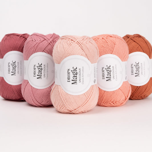 Fünf Knäuel der Wolle DROPS Magic in den Farben 79 dunkle rose, 22 zarte rose, 71 hell pfirsich, 54 pfirsich und 34 rostrot (von links nach rechts).