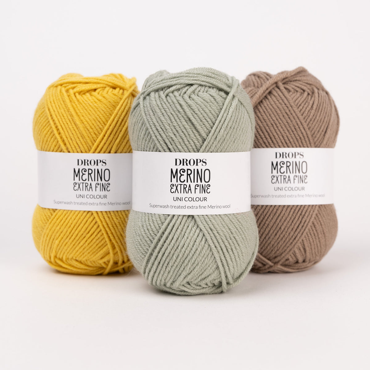 Drei Knäuel der Wolle DROPS Merino Extra Fine in den Farben 30 senf, 31 waldgrün und 51 café latte (von links nach rechts).
