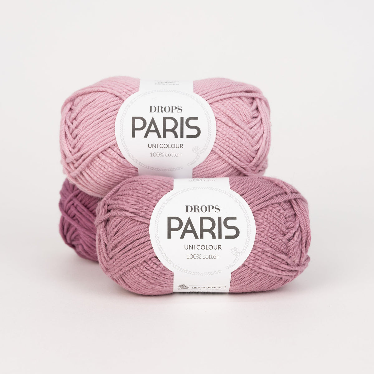 Drei Knäuel der Wolle DROPS Paris in den Farben 60 malve (hinten), 58 puderrosa (oben) und 64 amethyst (vorne).
