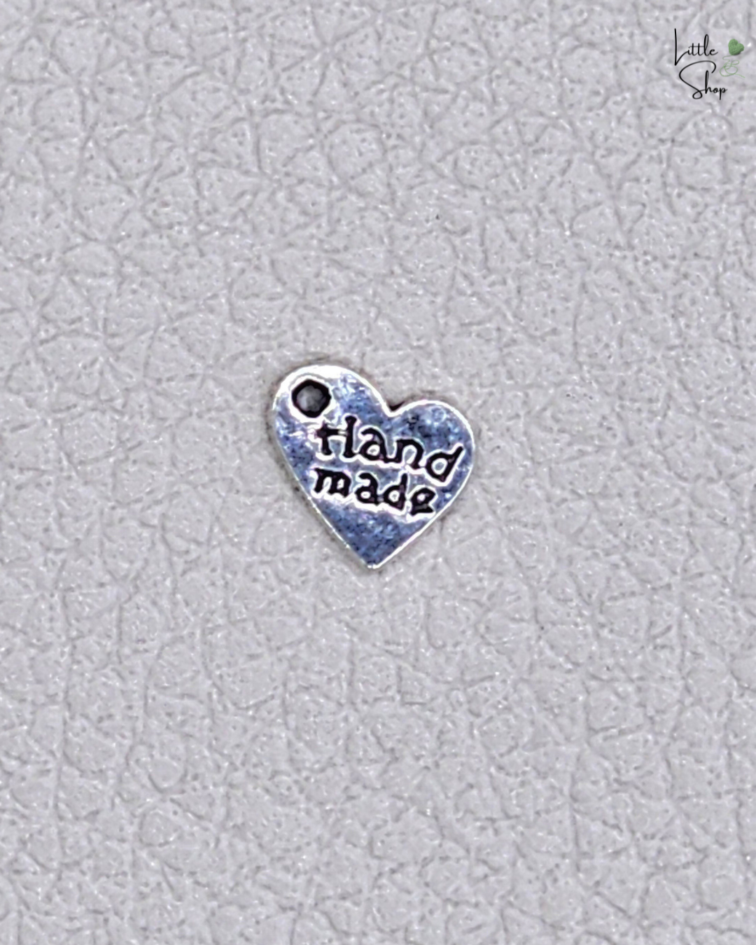 Silberfarbenes kleines Metallabel in Herzform mit dem Schriftzug Handmade und einer Ausstanzung, um das Label anzunähen oder aufzuhängen.